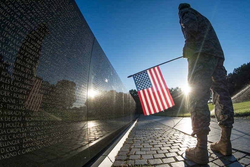 Soldier stands at Vietnam War Memorial with flag.